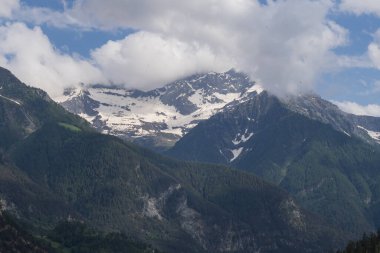 Alp dağlarının karlı sırtı bulutlu gün ışığı altında katmanlı yeşil orman tepelerinin üzerinde. Doğa ve macera temaları için güzel seyahat manzarası.