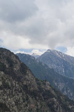 Karla kaplı zirveler, çarpıcı bulutlarla dolu yemyeşil orman yamaçlarının üzerinde duruyor. Seyahat, yürüyüş, irtifa ve manzara temaları için ideal barışçıl bir dağ manzarası..