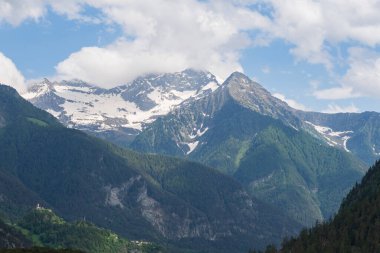 Karla kaplı zirveler, çarpıcı bulutlarla dolu yemyeşil orman yamaçlarının üzerinde duruyor. Seyahat, yürüyüş, irtifa ve manzara temaları için ideal barışçıl bir dağ manzarası..