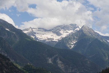 Karla kaplı zirveler, çarpıcı bulutlarla dolu yemyeşil orman yamaçlarının üzerinde duruyor. Seyahat, yürüyüş, irtifa ve manzara temaları için ideal barışçıl bir dağ manzarası..