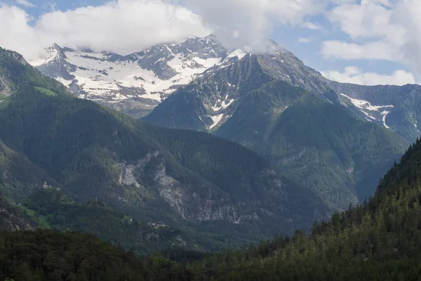 Karla kaplı zirveler, çarpıcı bulutlarla dolu yemyeşil orman yamaçlarının üzerinde duruyor. Seyahat, yürüyüş, irtifa ve manzara temaları için ideal barışçıl bir dağ manzarası..
