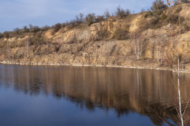 Terk edilmiş granit ocağında sakin bir göl. Kayalık kıyılar ve sonbahar ormanı sessiz sulara yansıyor. Gün batımında huzurlu doğal manzara.