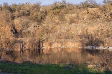 Terk edilmiş granit ocağında sakin bir göl. Kayalık kıyılar ve sonbahar ormanı sessiz sulara yansıyor. Gün batımında huzurlu doğal manzara.