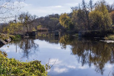 Durgun su, ağaç yansımaları ve eski taş sütunlarla doğal sonbahar nehri sahnesi. Yumuşak ışık ve mevsimsel renklerle sakin kırsal alan.
