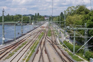 Güneşli bir günde göl kıyısını takip eden demiryolu hatları, kablolar, çakıl yatakları ve yeşil yaz bitkileri.