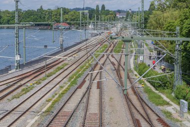 Güneşli bir günde göl kıyısını takip eden demiryolu hatları, kablolar, çakıl yatakları ve yeşil yaz bitkileri.