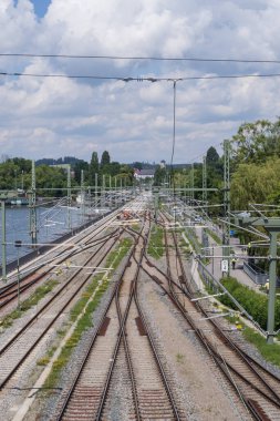 Güneşli bir günde göl kıyısını takip eden demiryolu hatları, kablolar, çakıl yatakları ve yeşil yaz bitkileri.