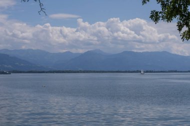 Lindau yakınlarındaki sakin Constance Gölü 'nde mavi su, uzak dağ sırtları, yumuşak bulutlar ve ufukta birkaç yelkenli tekne.