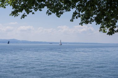 Lindau yakınlarındaki sakin Constance Gölü 'nde mavi su, uzak dağ sırtları, yumuşak bulutlar ve ufukta birkaç yelkenli tekne.