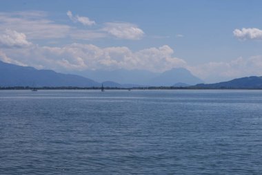 Lindau yakınlarındaki sakin Constance Gölü 'nde mavi su, uzak dağ sırtları, yumuşak bulutlar ve ufukta birkaç yelkenli tekne.