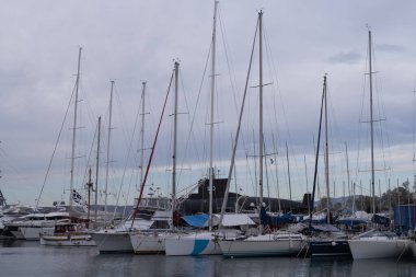 Yunanistan 'ın Atina kentindeki bir marinaya lüks yatlar ve tekneler yanaştı. Sakin deniz yüzeyi, modern liman altyapısını gösteren bulutlu gökyüzü altındaki gemileri yansıtır..