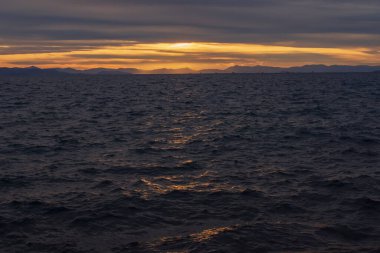 Akşam deniz manzarası, hareketli Ege Denizi dalgalarına altın gün batımı yansımalarını gösteriyor. Atina kıyısı yakınlarındaki sakin doğal manzara dramatik bulutlar ve sıcak ufuklar.