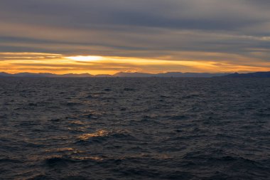 Akşam deniz manzarası, hareketli Ege Denizi dalgalarına altın gün batımı yansımalarını gösteriyor. Atina kıyısı yakınlarındaki sakin doğal manzara dramatik bulutlar ve sıcak ufuklar.