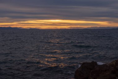 Akşam deniz manzarası, hareketli Ege Denizi dalgalarına altın gün batımı yansımalarını gösteriyor. Atina kıyısı yakınlarındaki sakin doğal manzara dramatik bulutlar ve sıcak ufuklar.