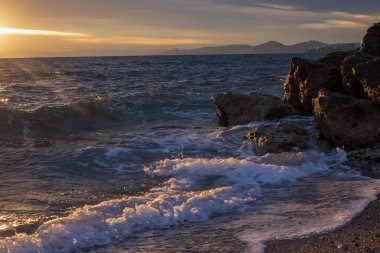 Gün batımında deniz manzarası, Yunanistan 'ın Atina kenti yakınlarındaki kayalık kıyılara vuran dalgaları gösteriyor. Sıcak güneş ışığı, desenli su yüzeyi ve uzak siluetler sakin bir Akdeniz akşam havası oluşturur..