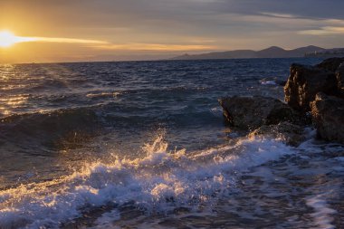 Gün batımında deniz manzarası, Yunanistan 'ın Atina kenti yakınlarındaki kayalık kıyılara vuran dalgaları gösteriyor. Sıcak güneş ışığı, desenli su yüzeyi ve uzak siluetler sakin bir Akdeniz akşam havası oluşturur..