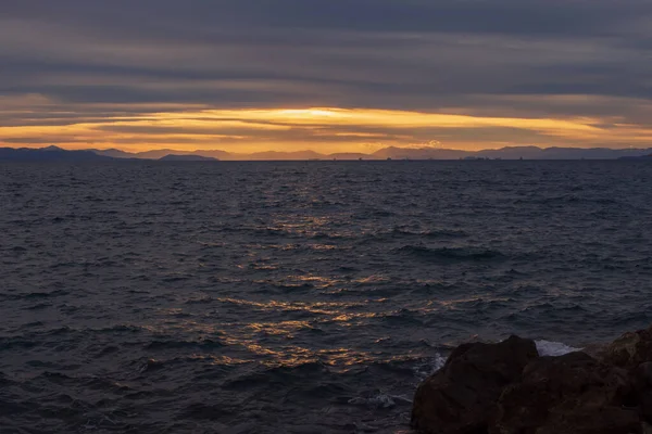 Akşam deniz manzarası, hareketli Ege Denizi dalgalarına altın gün batımı yansımalarını gösteriyor. Atina kıyısı yakınlarındaki sakin doğal manzara dramatik bulutlar ve sıcak ufuklar.