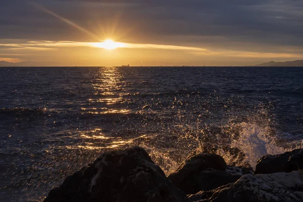 Gün batımında deniz manzarası, Yunanistan 'ın Atina kenti yakınlarındaki kayalık kıyılara vuran dalgaları gösteriyor. Sıcak güneş ışığı, desenli su yüzeyi ve uzak siluetler sakin bir Akdeniz akşam havası oluşturur..