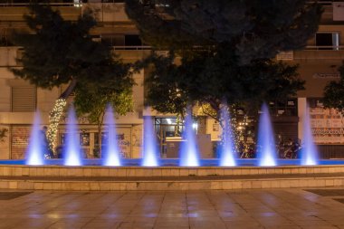 Yunanistan 'ın Atina kentinde gece vakti mavi ışıkla aydınlatılan şehir çeşmesi. Kamu meydanında dekoratif su jetleri, ağaçlar, sokak lambaları ve şehir akşam ortamı.