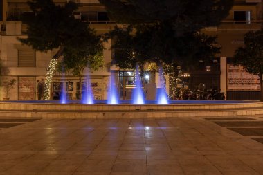 Yunanistan 'ın Atina kentinde gece vakti mavi ışıkla aydınlatılan şehir çeşmesi. Kamu meydanında dekoratif su jetleri, ağaçlar, sokak lambaları ve şehir akşam ortamı.