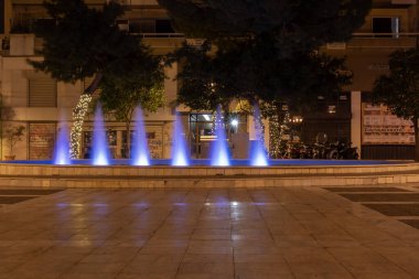 Yunanistan 'ın Atina kentinde gece vakti mavi ışıkla aydınlatılan şehir çeşmesi. Kamu meydanında dekoratif su jetleri, ağaçlar, sokak lambaları ve şehir akşam ortamı.