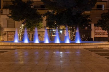 Yunanistan 'ın Atina kentinde gece vakti mavi ışıkla aydınlatılan şehir çeşmesi. Kamu meydanında dekoratif su jetleri, ağaçlar, sokak lambaları ve şehir akşam ortamı.