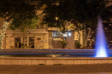Yunanistan 'ın Atina kentinde gece vakti mavi ışıkla aydınlatılan şehir çeşmesi. Kamu meydanında dekoratif su jetleri, ağaçlar, sokak lambaları ve şehir akşam ortamı.