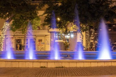 Yunanistan 'ın Atina kentinde gece vakti mavi ışıkla aydınlatılan şehir çeşmesi. Kamu meydanında dekoratif su jetleri, ağaçlar, sokak lambaları ve şehir akşam ortamı.