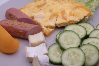 Sağlıklı kahvaltı tabağı omlet, dilimlenmiş sosis, peynir, salatalık ve marul. Basit ev yemekleri, dengeli beslenme ve günlük yiyecek konsepti..