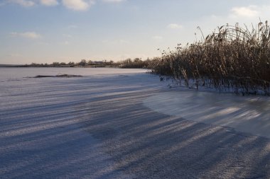 Kar ve kuru sazlıklarla kaplı donmuş bir nehrin üzerinde kış günbatımı. Alçak güneş uzun gölgeler ve yumuşak ışık yaratır. Sakin atmosfer, soğuk hava ve dokunulmamış doğa ifade eder..