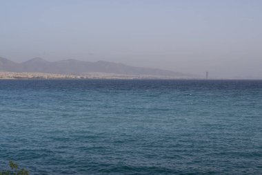 Açık mavi su, küçük kayalık ada ve kentin arkasındaki dağların yer aldığı Atina sahili manzarası. Doğal ışık, sakin yaz havası..