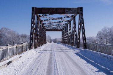 Buzlu yol kışın çelik bir köprüden geçiyor. Buz kaplı ağaçlar ve açık mavi gökyüzü arkaplanı.