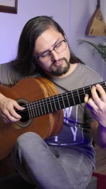 Bir kişi evde oturup klasik gitar çalar ve dikkatle panoya odaklanır. Sahne sıcak bir ışık, ahşap bir alet ve rahat bir atmosfer içeriyor.