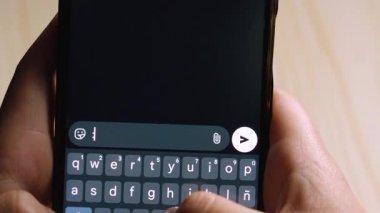 Simgeyi yazarken akıllı telefonu tutan ellerin yakın plan görüntüsü: Mesajlaşma uygulamasında D, karanlık temalı bir klavyede gönderme düğmesinin üzerinde gezinen bir başparmak.