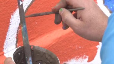 Bir ressamın elinin detaylı bir yakın çekimi fırçayı tutarken, yaratıcı duvar resimleri sırasında kalın ve canlı bir boyayı dokulu bir duvar yüzeyine titizlikle uyguluyor..