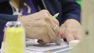 Yaşlı bir kadının ellerinin ön planda sarı bir makarayla çalışma tezgahındaki teknik düzeni ayarlamak için kalem ve silgi kullanışının detaylı görüntüsü.
