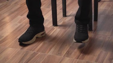 Oturan birinin bacakları ve ayakları siyah spor ayakkabı giyerek yakın plan çekimleri, ahşap zemin ile kapalı bir alanda hareket eden ritimik bir ayak oluşturur..
