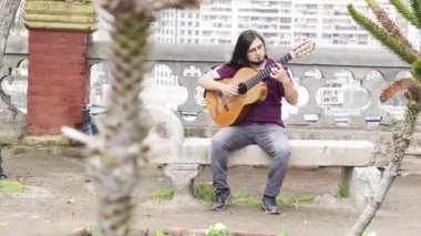 Uzun saçlı, keçi sakallı, mor gömlekli ve gri kotlu taş bir bankta oturan bir müzisyenin yatay geniş açılı görüntüsü. Odaklanmış bir ifadeyle klasik bir akustik gitar çalıyor..