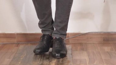 Kapalı bir yerde siyah deri dans ayakkabıları ve gri pantolon giyen, ahşap bir platformda flamenko dansı yapan birinin görüntüsü..