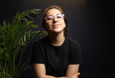 Yaratıcı stüdyodaki genç bir kadının profesyonel portresi yeşil bitkilerle dolu karanlık bir arka plana karşı. Modern yaşam tarzı, rahat duruş, doğal ifade, kopyalama alanı.