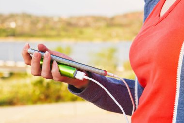 Bir kız dış güç bankasından şarj olurken açık havada akıllı telefon kullanıyor.