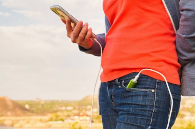 Bir kız dış güç bankasından şarj olurken açık havada akıllı telefon kullanıyor. Şarj aleti kotun cebinde.