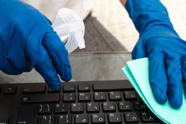 Mavi koruyucu eldivenli bir kız koronavirüsü, bakterileri, virüsleri ve mikropları önlemek için dezenfektanla klavyeyi temizliyor..