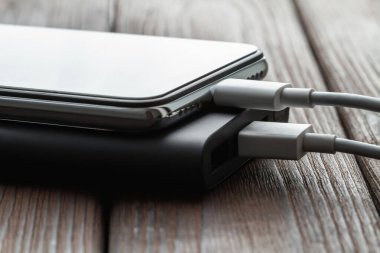 Akıllı telefon dış elektrik bankasından kabloyla kahverengi ahşap arka planda şarj oluyor. Yakın çekim, seçici odaklanma.
