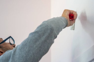 Gözlüklü bir adam kırmızı fırçalı beyaz bir iç duvar boyuyor, yakın görüş, günlük ev dekorasyonu, DIY yenileme konsepti