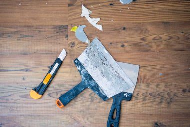 Alet bıçağı ve iki kullanılmış macun bıçağı tahta zeminde boya ve kağıt kırıntılarıyla dinleniyor. Ev yenileme ve onarım işlerini gösteriyor.