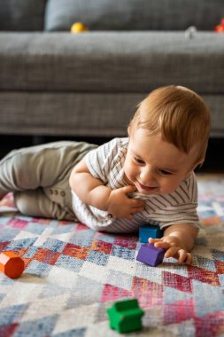 Gülen bebek desenli bir halıya uzanmış renkli ahşap bloklarla oynuyor, doğal ışıklandırma, rahat ev içi, erken gelişim konsepti