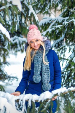 Güzel, gülümseyen beyaz bir kadının portresi. Kış ormanında duruyor. Mavi ceket ve pembe şapka giyiyor.