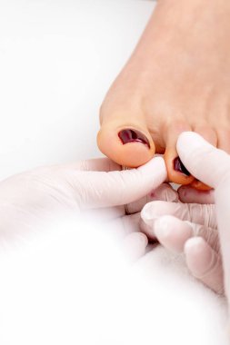 Manikür ustası, beyaz eldivenler giyen, kestane rengi ojeli kadın ayak tırnağı tutuyor.