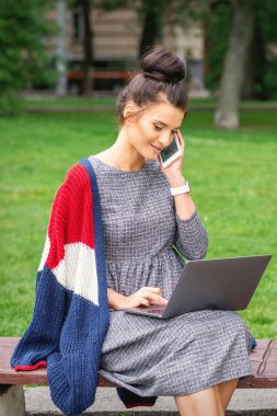 Öğrenci genç bayan akıllı telefonla konuşuyor. Parktaki bankta dizüstü bilgisayar kullanıyor.